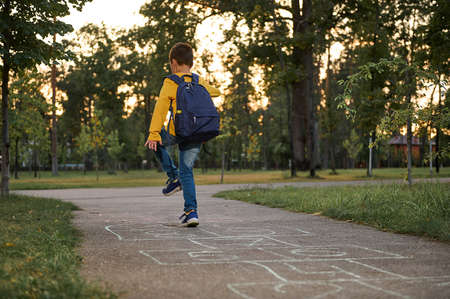 An active sporty boy with a school bag on his back plays hopscotch after school, takes turns jumping over the squares marked on the ground. Street children's games in classics.の写真素材