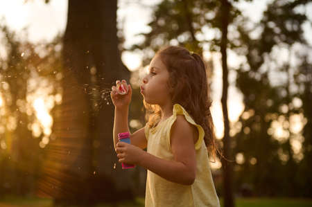 Beautiful gorgeous cute adorable little girl blowing soap bubbles at sunset. Carefree childhood, outdoors leisure activities. Baby girl playing games in the natureの写真素材