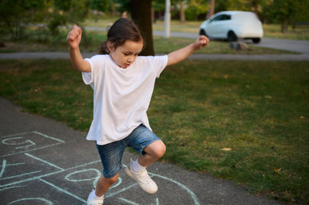 Street children's games in classics. Closeup of little Caucasian girl playing hopscotch on asphalt. Child playing hopscotch game on playground outdoors on a sunny day.の写真素材
