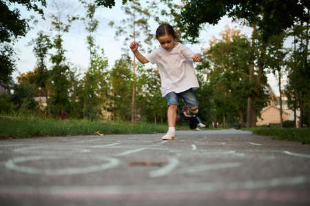 An active sporty 4 years old baby girl plays hopscotch , takes turns jumping over the squares marked on the ground. Street children's games in classics.の写真素材