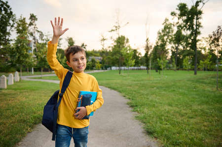 A charming handsome friendly schoolboy 9 years old in the park, with a backpack on one shoulder and notebooks in his hands, waves his hand, looks around and smiles sweetly with a toothy smile.の写真素材