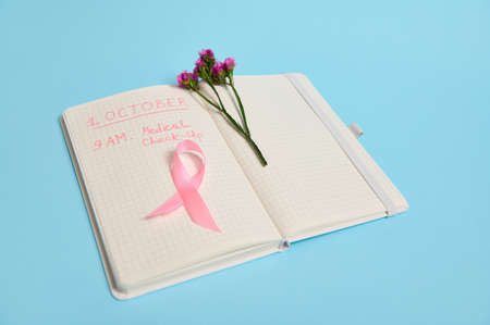 Flowers and pink ribbon, symbol of Breast Cancer Awareness Day, lying on a page with a line of notepad with inscriptions reminding of a medical check-up . October 1st, International Breast Cancer Dayの写真素材