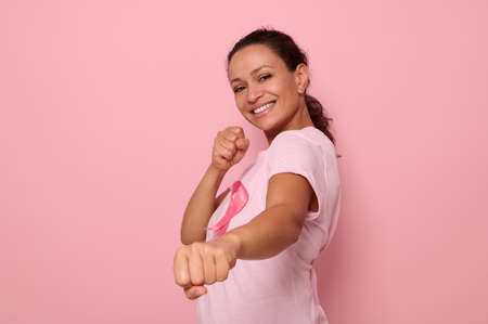 Pretty woman in pink t-shirt and cancer awareness ribbon stands in a fighting stance to mark the fight against cancer, in honor of October 1, smiles looking at camera, colored background, copy spaceの写真素材