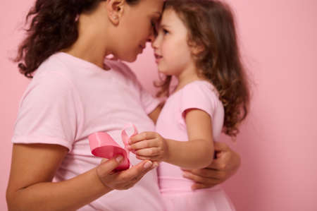 Focus on pink satin ribbon, symbol of International Breast Cancer Awareness Day, against blurred background of loving mother and lovely daughter. Female healthcare and medical education conceptの写真素材