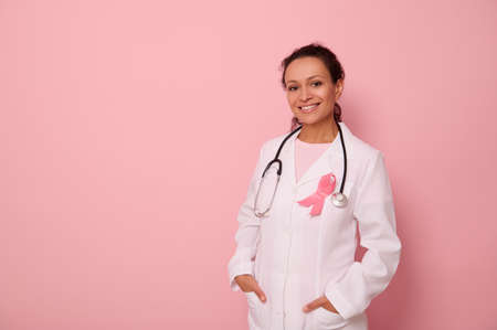 Portrait of African American womandoctor in medical coat and pink ribbon, stethoscope around neck, looking at camera, isolated on colored background, copy space. World Breast Cancer Awareness Dayの写真素材