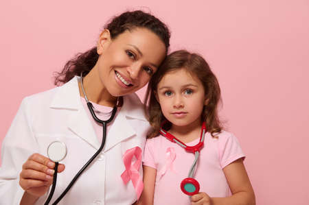Smiling female doctor and cute little girl, both with Breast Cancer Awareness pink ribbon and phonendoscope around their neck, showing stethoscope to camera, isolated on pink background, copy space.の写真素材
