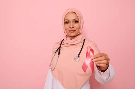 Gorgeous serene Arab Muslim female doctor in pink hijab holding Pink satin Ribbon, looking at camera, isolated on colored background, copy space. World Day of fight Breast Cancer, 1 st October conceptの写真素材