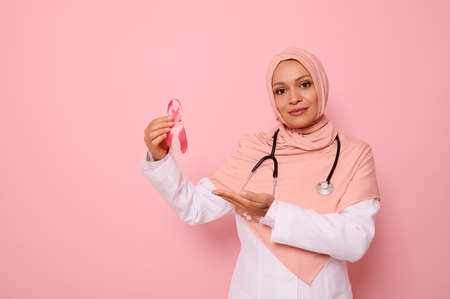 Confident Arab Muslim woman doctor in pink hijab and medical coat, showing pink Cancer Awareness ribbon, looking at camera. Pink background, copy space. International Day of fight oncological illnessの写真素材