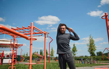 Handsome young adult 40 years old European sporty man stretching arms behind his back before training on the sportsground in open air. Mature sportsman enjoying workout outdoor on beautiful sunny dayの写真素材