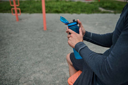 Close-up, cropped view of an unrecognizable athlete holding a fresh water bottle rehydrating his...