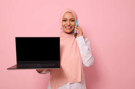 Attractive successful Arab Muslim woman in pink hijab talks on mobile phone, smiles with toothy...