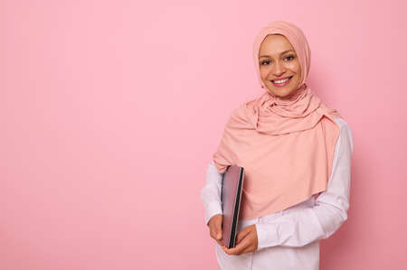 Gorgeous Muslim woman in stylish traditional religious outfit with covered head in pink hijab smiling with toothy smile posing against colored background with a laptop in hands. Copy spaceの写真素材