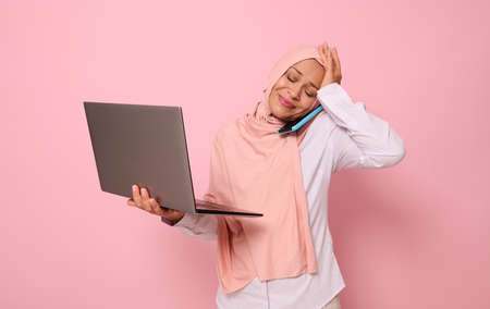 Puzzled Muslim woman in hijab, with laptop, talking on the phone, holding her head because of multitasking. Concept of a busy young Arab business lady, freelancer, office worker on colored backgroundの写真素材