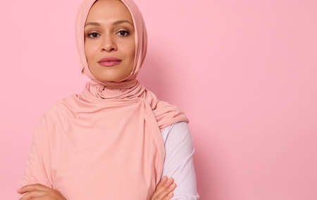 Headshot, close-up face portrait of a gorgeous Arabic Muslim woman in pink hijab posing looking to camera with attractive gaze, confident look and crossed arms, on pink background with copy spaceの写真素材