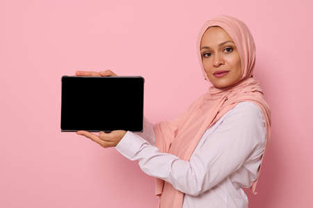 Confident Muslim Arab beautiful woman with covered head in hijab holds horizontally a digital tablet and shows its blank screen looking at the camera, isolated on pink background with copy spaceの写真素材