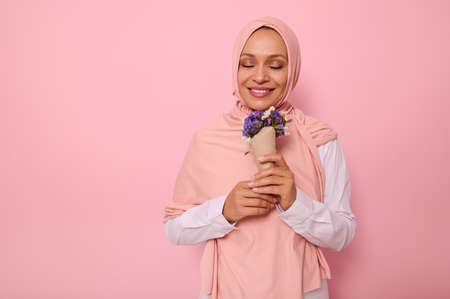 Portrait on pink background of beautiful Muslim Arab woman in hijab with closed eyes and charming smile enjoying the smelling of meadow flowers wrapped in craft paper. International Women's Day conceptの写真素材