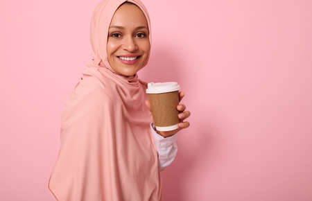 Close-up. Muslim Arabic woman with covered head in hijab holds disposable cardboard takeaway cup, smiles toothy smile, looking at camera, standing three quarters against colored background, copy spaceの写真素材