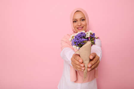 Focus on a beautiful simple bouquet of meadow wildflowers in purple tones, wrapped in brown craft paper in the hands of an attractive smiling Muslim woman wearing a hijab. Pink background copy spaceの写真素材