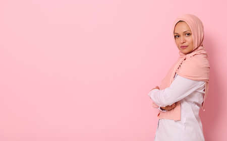 Confident portrait of Arabic muslim beautiful woman with attractive look and gaze, wearing pink hijab and standing sideways against colored background with copy spaceの写真素材