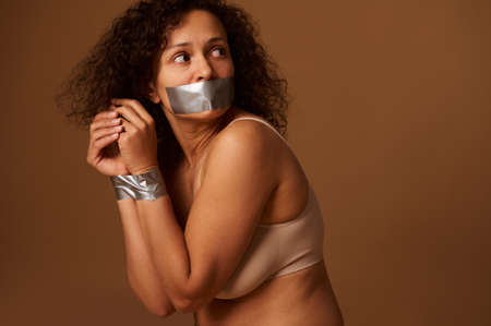 Close up portrait of frightened desperate woman with her mouth closed and her hands tied, looking back with hope and praying for help in ending violence against women. Social concept, dark background.の写真素材