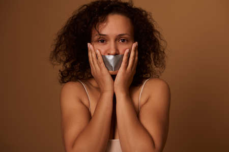 Frightened mixed race woman with sealed mouth looks desperately at camera, holding her hands on her cheeks, isolated on dark colored background with copy space. Concept of STOP violence against womenの写真素材