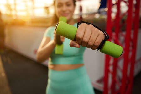 Soft focus on a sporty woman's outstretched hand holding a small dumbbell, standing on the background of a boxing arena. Blurred fitness woman exercises outdoors at sunriseの写真素材
