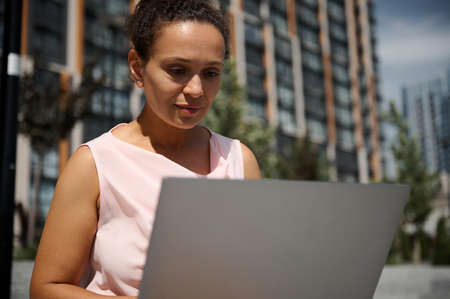 Close-up portrait of middle aged African or Latin American woman, business lady, office manager, worker, employee in casual attire working on laptop on the high buildings backgroundの写真素材