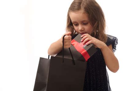 Concept for Black Friday, isolated portrait of an adorable baby girl in dark blue evening dress putting a gift with red ribbon into a black packet, copy space. Shopping, sales and purchases concept.の写真素材