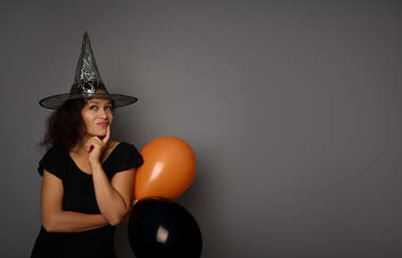 Beautiful mysterious pensive young mixed race woman in black and wizard hat, looking up thoughtfully , posing against a gray background with copy space for ad. Halloween party conceptの写真素材