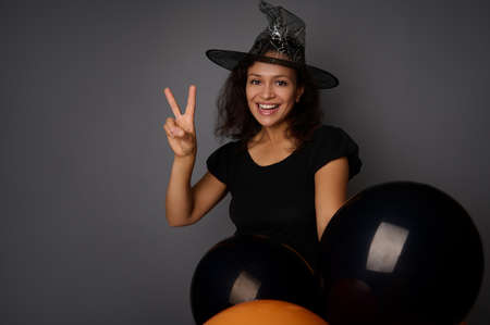Happy smiling toothy smile Hispanic woman wearing wizard hat, dressed in witch carnival costume for Halloween party, shows a peace sign, poses against gray background with black balloons, copy spaceの写真素材