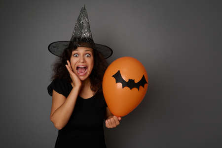 Scared woman in carnival witch costume and wizard hat, screams, holds an orange colorful balloon and puts her hand on her cheek, looking at camera. Halloween concept on gray background with copy spaceの写真素材
