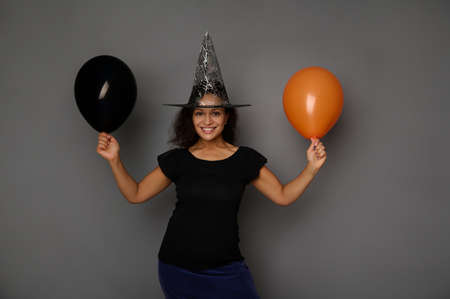 Beautiful attractive woman dressed in witch black costume and wizard hat, stands against gray background with orange and black air balloon, smiles looking at camera. Halloween concept with copy spaceの写真素材