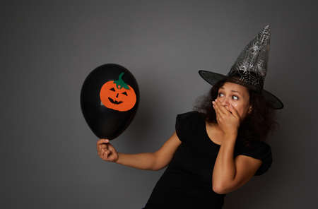A stupefied woman dressed in black witch carnival attire covers her mouth from horror, looks at a black air balloon with felt-cut pumpkin in hand. Halloween concept with copy space on gray background.の写真素材