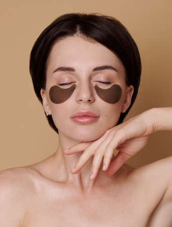 Young woman with healthy fresh clean skin poses on beige background , reduces under eye puffiness, dark circles, applies moisturizing patches under eyes, cares of eye skin beauty. Wellness spa conceptの写真素材