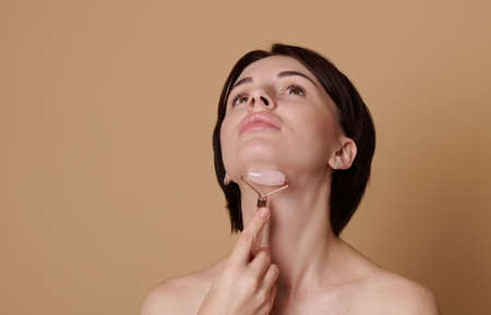 Young brunette woman doing a lifting massage on her neck, using a jade roller massager. Beauty portrait on beige background with space for ad.の写真素材