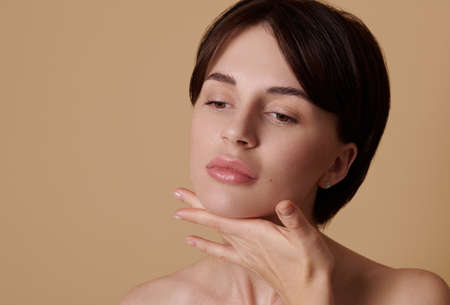 Close-up beauty portrait on beige colored background of attractive young European woman doing anti-aging face and neck massage. Skin and body care, wellness and wellbeing concept, copy space for adの写真素材