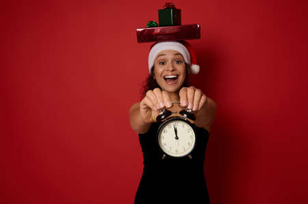 Gorgeous woman in black evening dress and Santa hat, puts gift boxes on her head, smiles toothy smile and shows alarm clock with time around 12 o'clock at night, looking at camera. Christmas conceptの写真素材