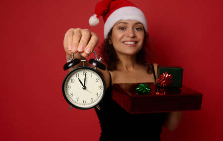 Focus on alarm clock in the hand of blurred cheerful woman in Santa hat, smiling with beautiful toothy smile, holding Christmas gift boxes wrapped in glitter red green gift paper. Copy space for adの写真素材