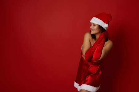 Cute mixed race woman wearing Santa hat, dress and red satin gloves, holds her hands near her cheek, smiles toothy smile, posing over red colored background with copy space for Christmas advertising.の写真素材