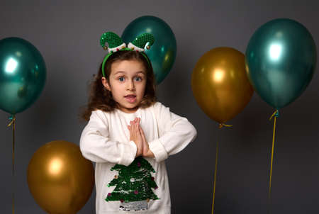 Pretty little girl wearing Christmas sweater and elf hoop poses with hands palm to palm, looks at camera, isolated over gray background with golden metallic green air balloons with copy space for adの写真素材