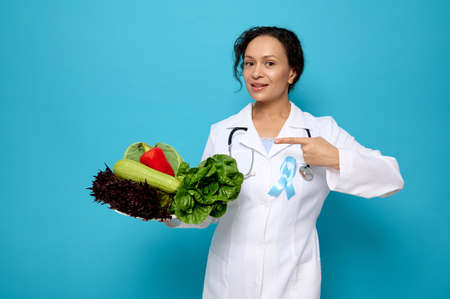 Beautiful woman, female doctor nutritionist wearing a white medical gown with blue awareness ribbon points on a plate full of healthy raw vegan eating. World Diabetes Day concept with copy spaceの写真素材