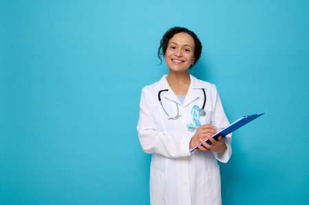 Middle aged mixed race doctor in white medical gown, wears a blue awareness ribbon, poses with a clipboard in her hands, cutely smiled looking at camera,copy space. World Diabetes Awareness Dayの写真素材