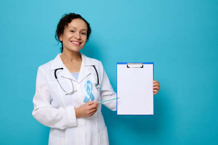 Gorgeous female doctor in white medical gown and blue awareness ribbon smiles toothy smile looking at camera and shows a copy space on empty blank paper sheet on clipboard. World Diabetes Day conceptの写真素材