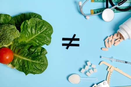 Concept for World Diabetes Awareness Day. Vegan food, leaves, salads, vegetables, inequality sign and medicines, pharmaceutical pills, sugar cubes, insulin syringe and stethoscope on blue background.の写真素材