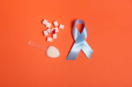 Flat lay of pure refined white sugar cubes with blood drop and blue satin ribbon, symbol of World diabetes awareness day 14 November. Isolated over colored orange background, copy space for medical adの写真素材