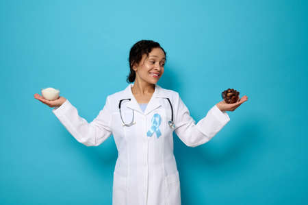 Sugar and diabetes concepts for World Diabetes Awareness Day. Smiling nutritionist in medical gown an blue ribbon holding a bowl with refined white sugar in one hand and ripe sweet dates in the otherの写真素材