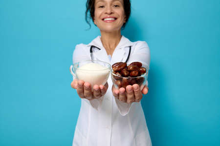 World diabetes Day. Focus on transparent glass bowls with ripe sweet dates and unhealthy white refined sugar on the hands of female doctor nutritionist, isolated over blue background with copy space.の写真素材