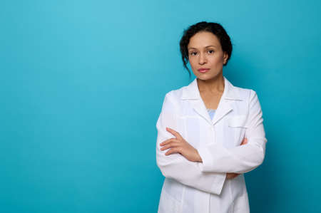 Confident portrait of beautiful serene woman doctor in white medical gown, looking at camera posing with crossed arms against blue colored background with copy space for medical advertisingの写真素材