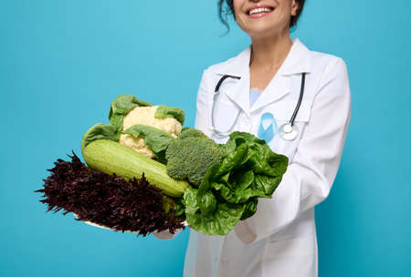 Raw vegetables, healthy vegan food, greens and salads in the hands of smiling nutritionist wearing a medical gown with blue diabetes awareness ribbon, isolated over colored background, copy space.の写真素材