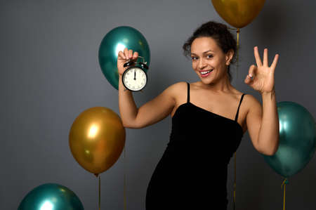 Cheerful happy mixed race woman holds alarm clock, it's midnight, showing OK sign smiles looking at camera, isolated on gray background with balloons. New year, Merry Christmas concept for advertisingの写真素材
